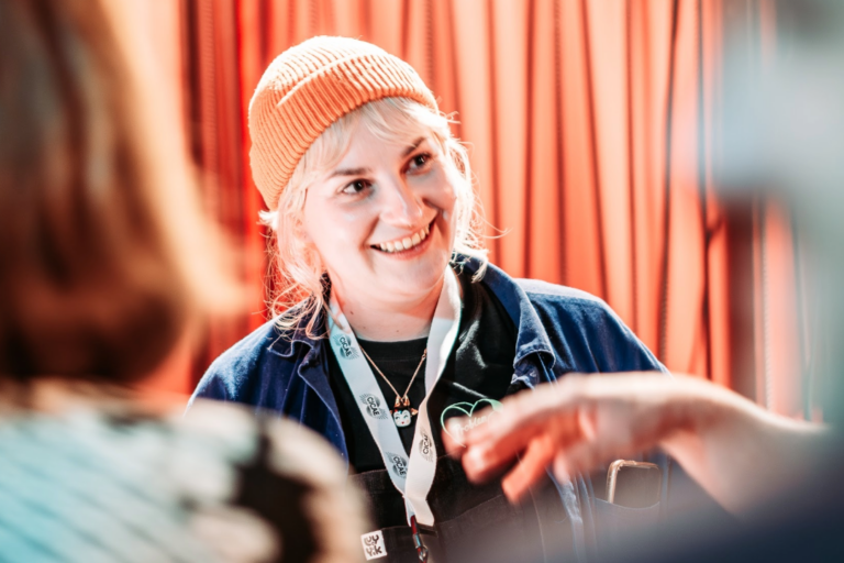 A woman with blonde hair and an orange beanie is smiling against an orange curtain in the background.