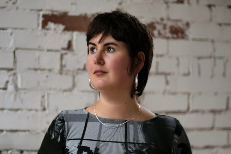A person with shoulder-length brown hair, silver hoop earrings and a silver necklace looks to the right, over an exposed white brick background.